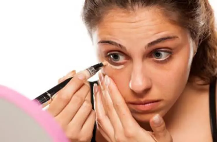 A young lady applying concealer below one of her eyes