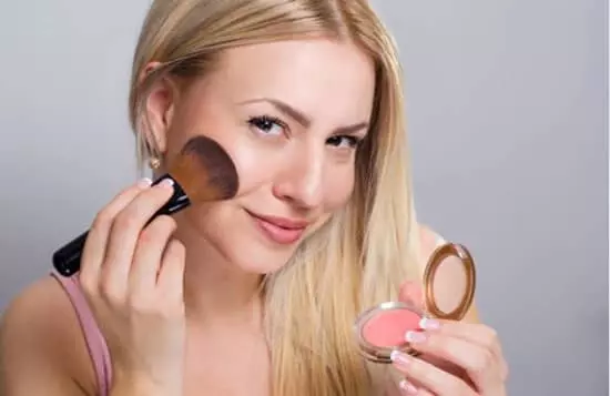 A young lady looking in mirror while applying a light pink blush to the apples of her cheeks using a blush brush and pressed compact with mirror