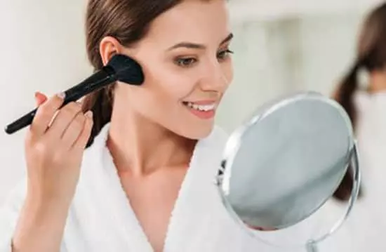 A young woman expertly applying bronzer to her face with a brush while looking into a mirror enhancing her features with precision and skill