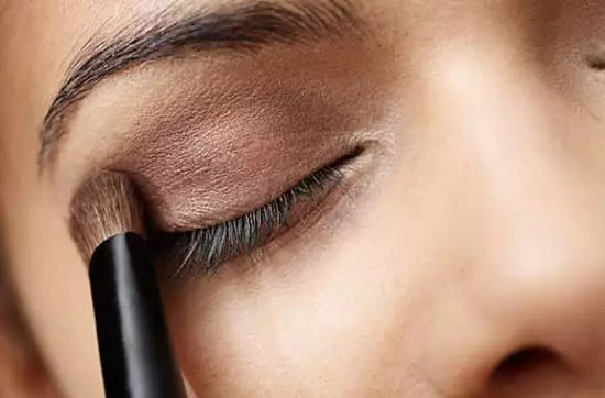 A lady applying eyeshadow to the top of her right eyelid with an eyeshadow brush