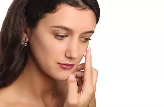 A young woman gently applying face primer with her finger to her left cheek with tenderness and care