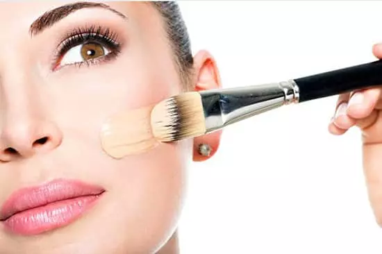 A pretty young lady applying foundation to the left cheek of her face with a foundation brush