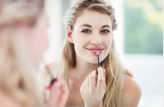 A happy young lady applying lipgloss to her lower lip while looking in the mirror