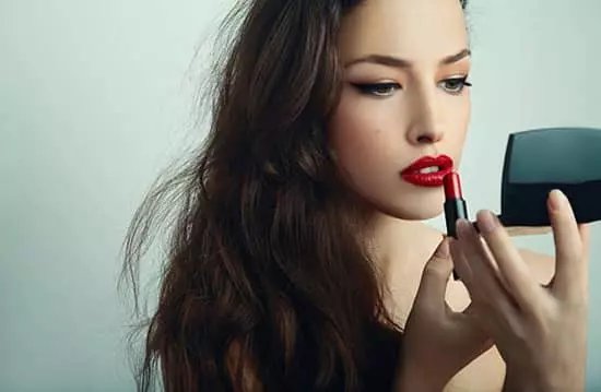 A young lady applying red lipstick to her upper lip while looking into a mirror