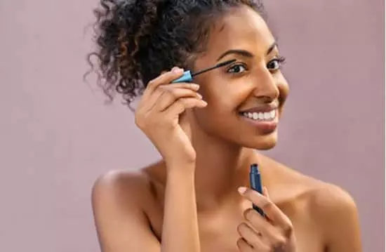 A smiling young woman applying mascara makeup with a wand to the base of her upper eyelashes