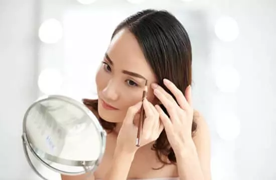 An attractive young asian woman skillfully using an eyebrow pen to enhance her brows and natural beauty while looking into a mirror in a bathroom