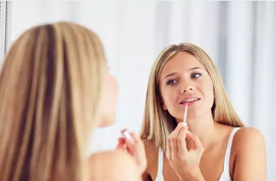 A young woman applying lipgloss to her upper lip while looking into a mirror